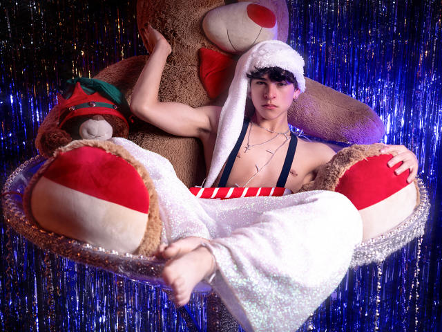 LukeHowlandy with muscular arms, wearing a white fluffy hat, striped underwear, and glittery pants, sits in a giant stuffed animal chair, surrounded by large red and white pillows.
