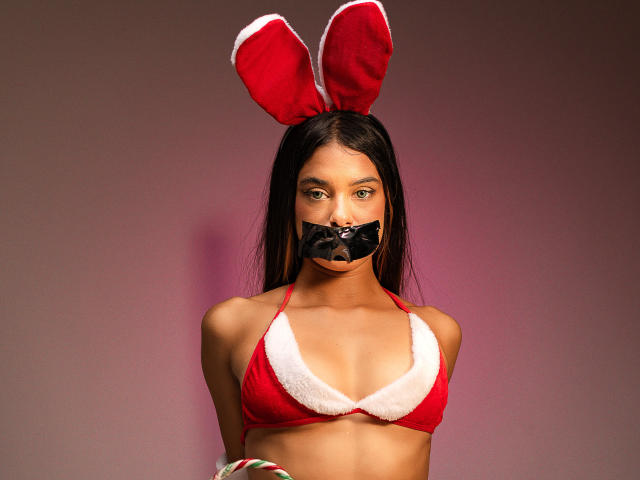 VanelopeRusso with dark hair, red bra with white fur trim, black tape over mouth, red bunny ears, brown skin, and green eyes, looking directly at camera.