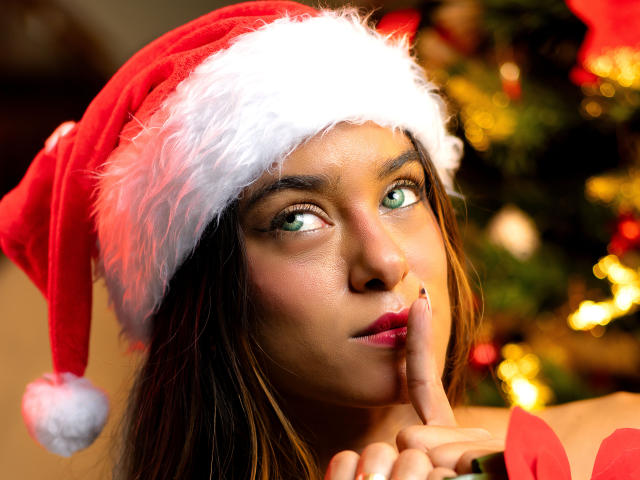 VanelopeRusso with green eyes, wearing a Santa hat, has her finger to her lips, looking slightly to the side, showing off her glossy red lips and a cute, sexy face.