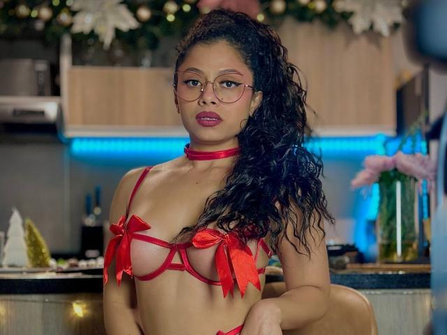 IvannaAndFriend in red lingerie with ribbons covering her tits, wearing a choker, glasses, and curly black hair, in a kitchen with Christmas decorations.