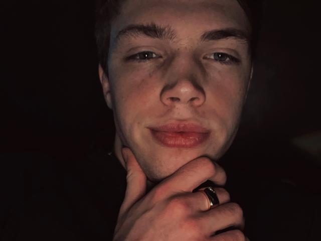 SashaMakedonski has light skin, blue eyes, and a slight smile. His hand with a black ring rests on his chin. Dark background, subtle light on his face.