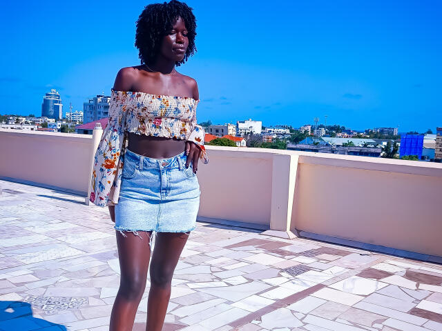 KharlaQueen stands on a rooftop with a clear blue sky. She has dark skin and curly black hair. She wears an off-the-shoulder, floral crop top and high-waisted, light blue denim skirt. Her top's sleeves are long and slightly puffed. She has a slim, athletic build. The rooftop has a patterned, light-colored floor. The cityscape in the background is blurred. The image is brightly colored with a vivid blue sky.