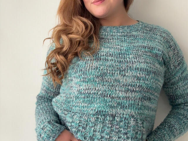 LaurenSpitz with wavy brown hair, wearing a green and blue textured knitted sweater, her tits subtly visible, standing against a plain white wall.