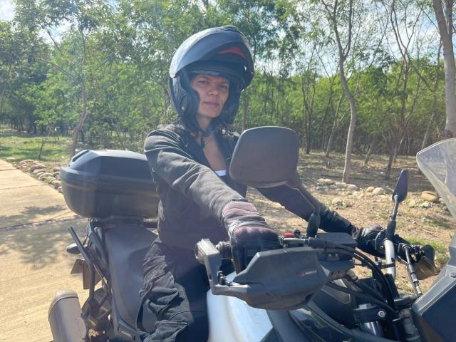 DulceDanesa with black helmet, wearing dark jacket, gloves, and black pants, riding a black scooter, sunlight, trees, and a concrete path in the background.