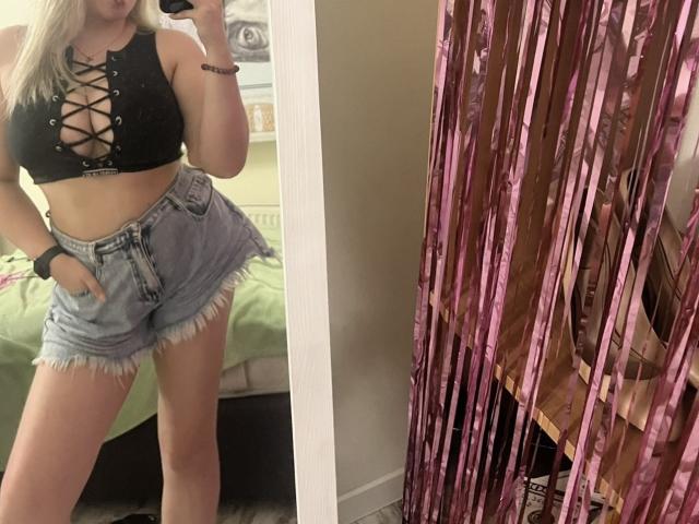 Een vrouw met blond haar, een zwart leren topje met veters, waarbij haar borsten zichtbaar zijn, en hoge, versleten denim shorts, die een spiegel-selfie maakt, en een zwarse armband en polshorloge draagt..