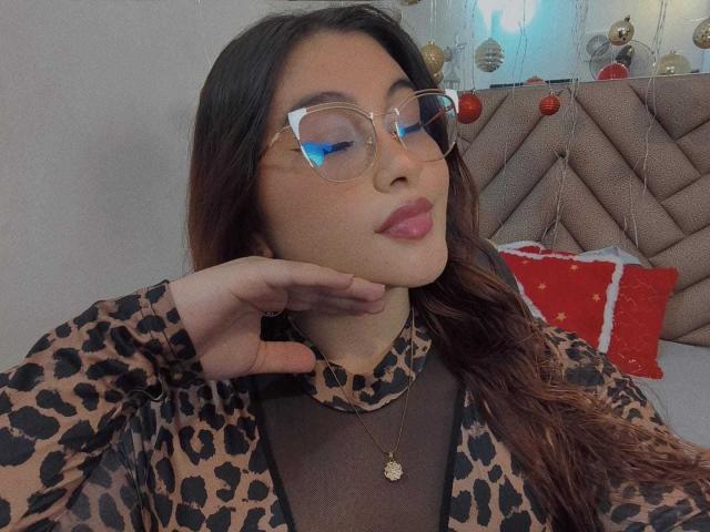 HelueJamil with glasses, leopard print top, and pink lipstick, resting chin on hand, brown wavy hair, gold necklace, looking at phone, subtle reflection in glasses.