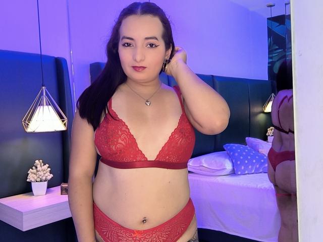 AnnaLeah with red lace bra and matching panties, showing her tits and ass, has a belly button piercing, dark hair, and a confident smile, standing in a bedroom.