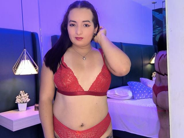 AnnaLeah stands in a bedroom wearing red lace lingerie, showing her boobs and pussy, with a hand in her dark hair, a small belly piercing, and a calm expression.