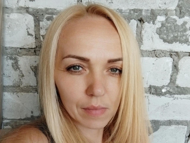 SallySweet-hot has blonde hair, blue eyes, and a slightly pouty face with subtle makeup, showing her tits in the image. She's against a white brick wall.