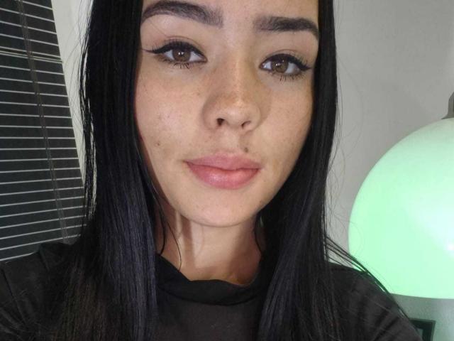 EvelynHartmann has long black hair, brown eyes, and full pink lips with a slight smile. She's wearing black eyeliner and a sheer black top. Her face is freckled.