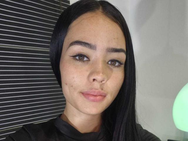 EvelynHartmann has straight black hair, brown eyes, and a light brown skin tone with small freckles. She's wearing black eyeliner and a black shirt. Her ass is not visible.