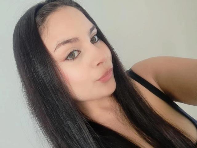 CharlotteTeylor with long black hair, green eyes, and subtle makeup, wearing a black top, showing her tits slightly. She has a cute, pouty face and a serious expression.