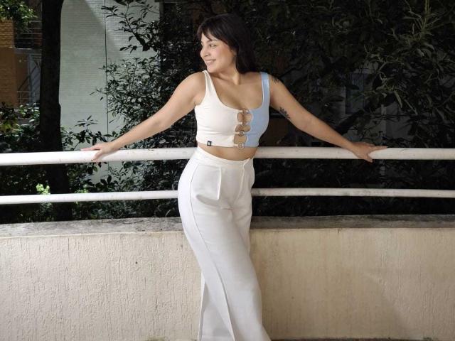 CharlotteTeylor with small tits in a white crop top, white high-waisted pants, standing with arms on a railing, smiling, dark hair, tattoos on her left arm, daytime.