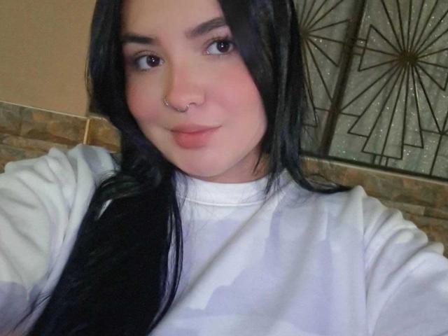 ValeriJoness with black hair, nose ring, and white shirt, smiling slightly, showing light pink lips, with soft pink cheeks, and brown eyes. Her tits are not visible.