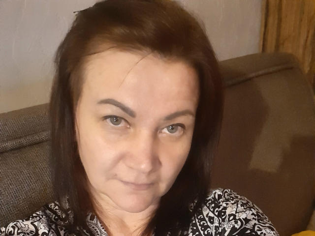 Close-up of CassieStefana, a middle-aged woman with light skin and brown hair, slightly parted to the right. She has small, dark eyebrows and brown eyes. Her expression is neutral, with a slight, closed-mouth smile. She's wearing a black and white patterned shirt. The brown sofa behind her is textured. The light is soft, creating mild shadows on her face. The image is sharply focused, emphasizing her facial features.