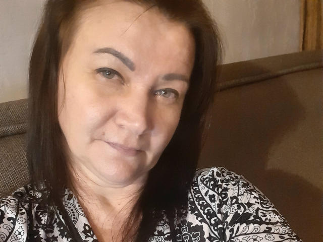 CassieStefana has brown hair, blue eyes, and a slight smile, wearing a black and white patterned shirt, looking directly at the camera. She's sitting on a brown couch.
