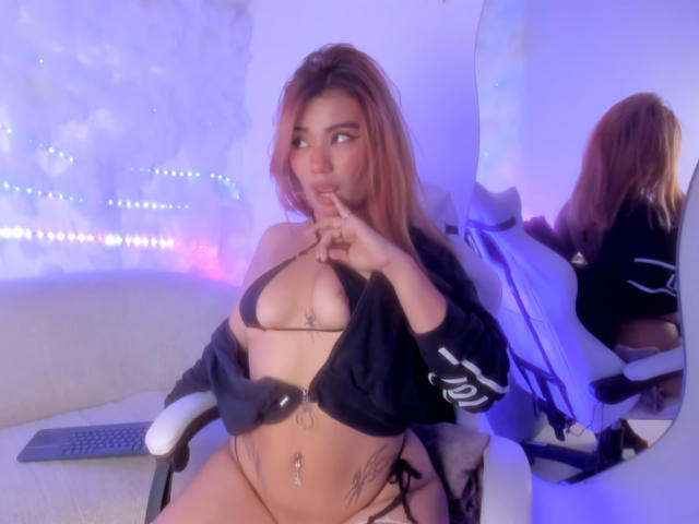 LiahMillerr with brown hair, wearing a black bikini, showing her small tits and pierced belly button, finger in mouth, sitting on a chair, reflection in mirror.