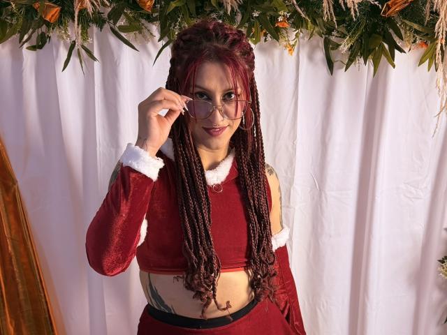 SofiFw with red braids, wearing a red velvet Santa crop top with white fur trim, holding glasses, showing a tattoo on her side, smiling, in a festive setting.