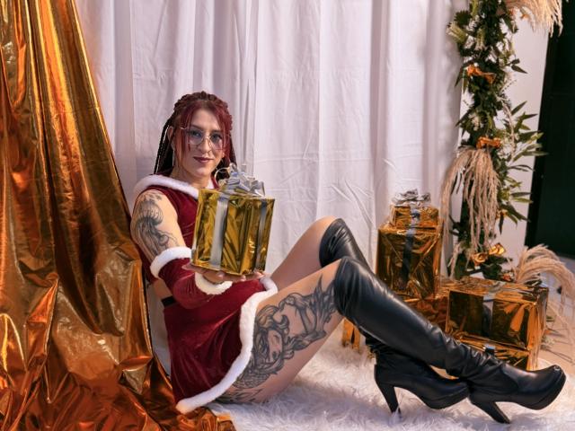 SofiFw in a red velvet Santa dress with white fur trim, black thigh-high boots, holding a gold gift box, showing tattoos on her thigh and arm, smiling.