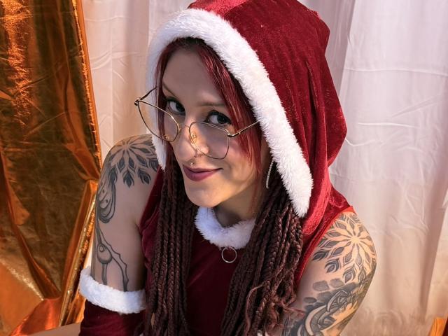 SofiFw with red braids, wearing a Santa hat, glasses, and tattoos on arms, smiling at camera, showing off tits with red velvet dress, white fur trim.