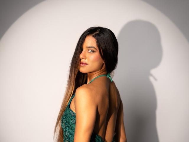 VanelopeRusso with long, straight brown hair, green halter dress, backless, looking over shoulder, shadow on white wall, smiling, red lipstick, green eyes, smooth tan skin.