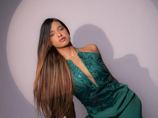 VanelopeRusso with long brown hair, green dress, deep V-neck, and sequins, leaning against a wall, shadow on her face, looking slightly to the side, with a soft expression.