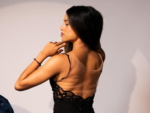 VanelopeRusso with dark hair, wearing a black lace dress, showing her back, fingers near her chin, tan skin, slim build, subtle makeup, and a black bracelet.