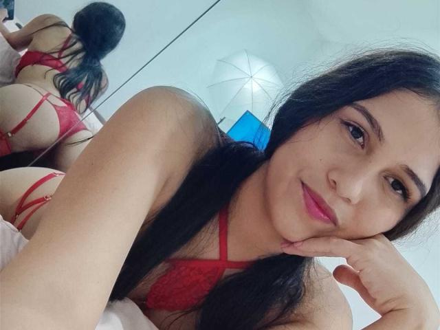 ValentinaMoreau with long black hair, red lace bra and thong, lying on bed, looking at camera, smiling, showing her ass in mirror reflection, pink lips, brown eyes.