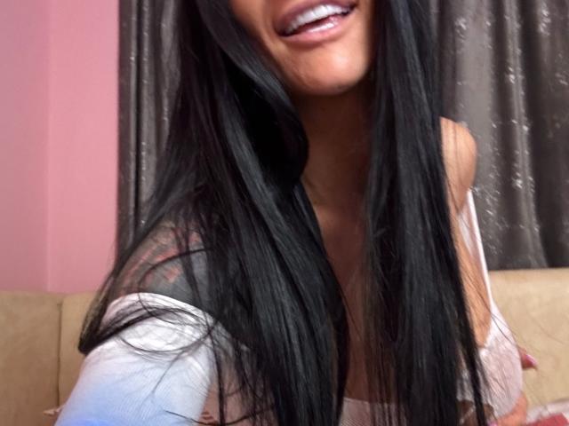 AndraD has long black hair, a smiling face with white teeth, and is wearing a pink top that shows her tits. Her top is off her shoulders.