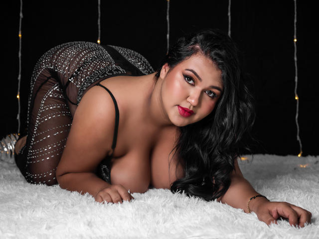 DojaAissha, curvy, dark-haired, wearing a sheer black dress with sparkles, on all fours, on white fur, exposing her large, round tits and slightly visible pussy, with red lips.