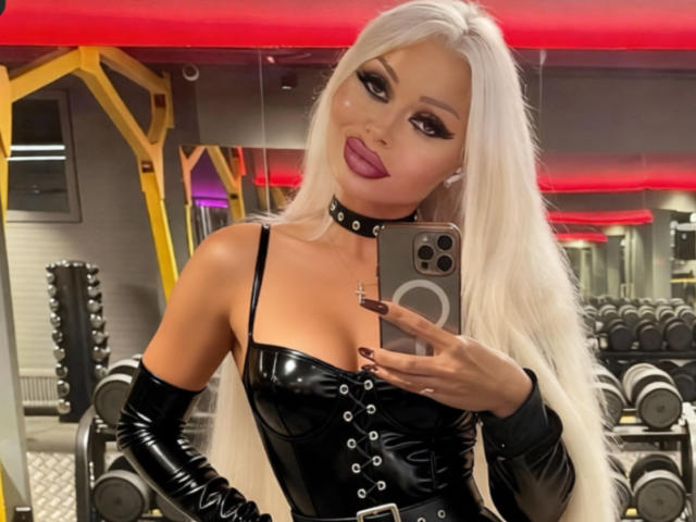 RoxyRoyale with long white hair, black latex top, choker, and gloves, taking a selfie in a gym, showing her tits and pouty lips, holding a phone.