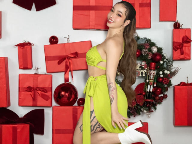 LilaOwens with long brown hair, green bikini top and skirt, white boots, red gifts, and Christmas decor, smiling, showing tattoo on thigh, and her ass.