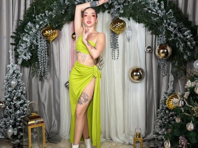 LilaOwens stands in a festive, green-wrapped setup. She's wearing a bright green, strapless, one-shoulder dress with a high slit on the left, exposing tattoos on her thigh and side. Her right arm is raised, holding a silver ornament. She has light skin, red lipstick, and her blonde hair is pulled back. The background features green wreaths with silver decorations and a gray curtain. She's wearing white socks. The image is highly detailed and colorful.