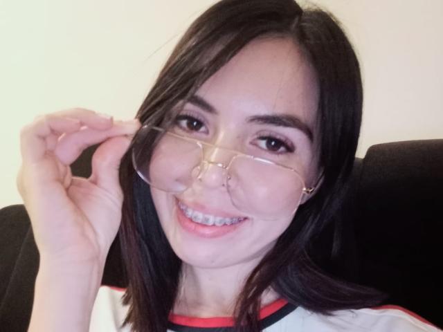 MollyTaylorx with black hair, clear glasses, smiling, holding glasses with right hand, wearing a white and red shirt, showing her teeth and looking at the camera.