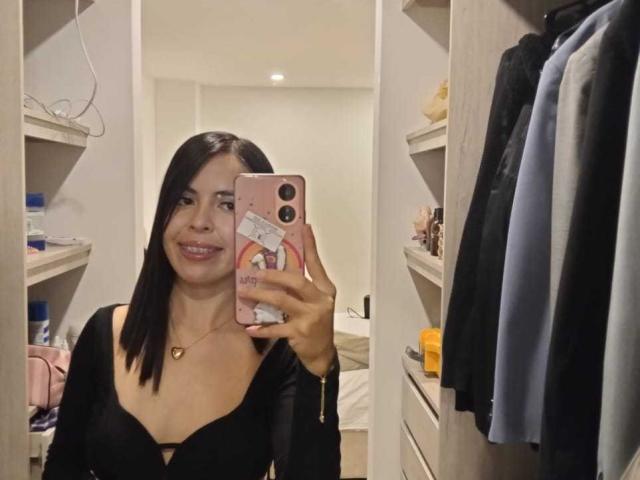 MollyTaylorx with black bob, smiling, wearing a low-cut black top, showing her tits, holding a pink phone, mirror selfie in a closet with clothes.