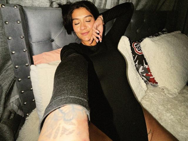 ThaliaAres with short black hair, eyes closed, wearing a black long-sleeve bodysuit, arms above head, legs spread, tattooed arm, lying on a gray tufted bed.