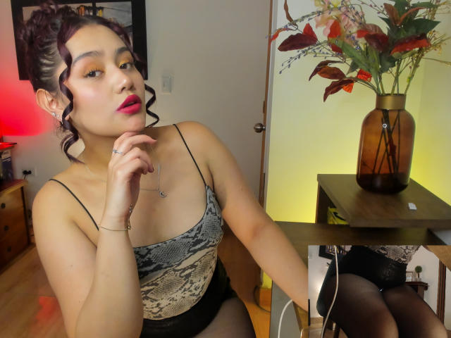 JessiicaBeltran with curly black hair, pink lipstick, snake-pattern camisole, black shorts, and black tights, hand on chin, red lamp glow, brown vase with flowers on table.