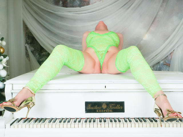SquirtyCumShow, Beine gespreizt, trägt neon-grüne Spitzendessous, hohe Strümpfe und goldene Absätze, auf einem weißen Klavier, Kopf nach hinten geneigt, freigelegte Vulva, sichtiges Gesäß..