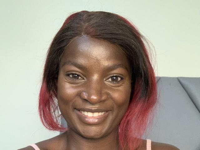 JennyNorah has dark brown skin, red and black hair, brown eyes, and a smile showing white teeth. She's wearing a pink top, looking happy and cute.