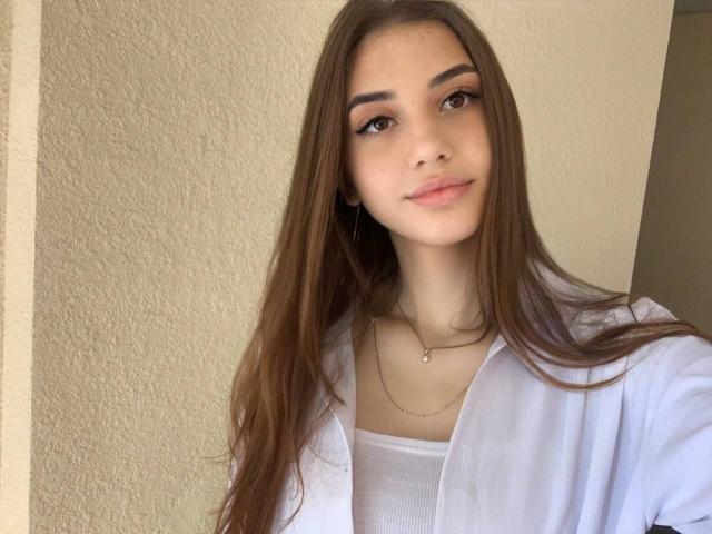GinnyQueen has long brown hair, brown eyes, and a subtle smile. She's wearing a white shirt over a white tank top, with a delicate necklace.