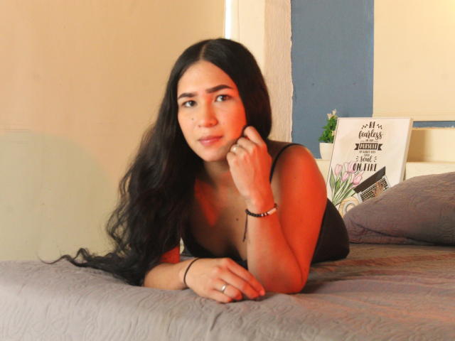 CelesteLeroy with long black hair, lying on a bed, wearing a black tank top, looking to the side, resting her chin on her hand, red light on her face.