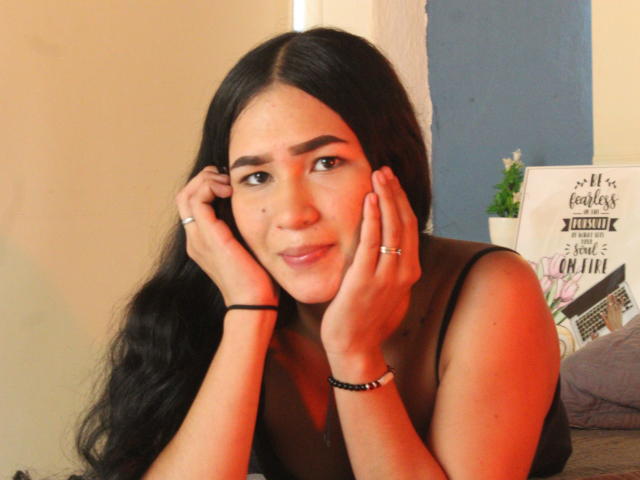 CelesteLeroy with long black hair, hands on her cheeks, slight smile, brown eyes, tan skin, small moles on her face, black sleeveless top, rings on fingers, bracelet on wrist.
