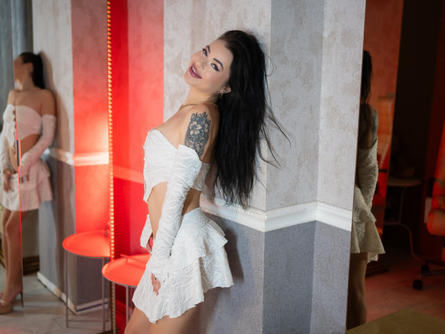 SelenaBellax with long black hair, white lace dress, and tattoos on her right shoulder, leaning against a wall, looking back with a smile, showing her tits and ass.
