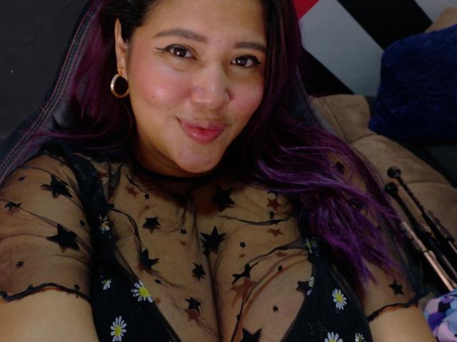 IamAhYours has medium brown skin, dark eyes with black eyeliner, and purple-tinted hair. She wears gold hoop earrings and a sheer black top with star and daisy patterns, revealing a black bra. Her top is slightly translucent, showing her cleavage. She has a small nose and a slight smile. The image is a close-up selfie. Her hair is straight and falls over her right shoulder. The lighting is even, highlighting her facial features and outfit. The texture of her top is delicate and patterned.