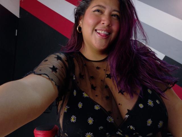 IamAhYours has long, purple-dyed hair and is smiling at the camera. She's wearing a black, sheer top with star patterns and small daisy prints, showing her large breasts. She has black earrings and is holding a red cup in the foreground. The image is a close-up selfie with a slightly low angle. Her skin is light brown, and she has a natural, confident look. The background has red, black, and white diagonal stripes.