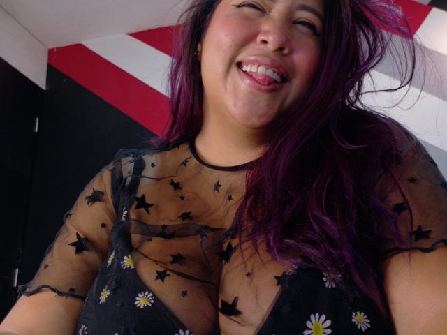 Close-up of IamAhYours with a wide, playful smile. She has long, wavy purple hair and is wearing a sheer black top with star and daisy patterns, exposing her large breasts. Her tongue is out, and she's looking slightly upward. The top is semi-transparent, revealing her skin underneath. The background has red and white diagonal stripes. The image is candid, with a casual, fun vibe. Her makeup is subtle, with black eyeliner. The focus is on her face and upper body.