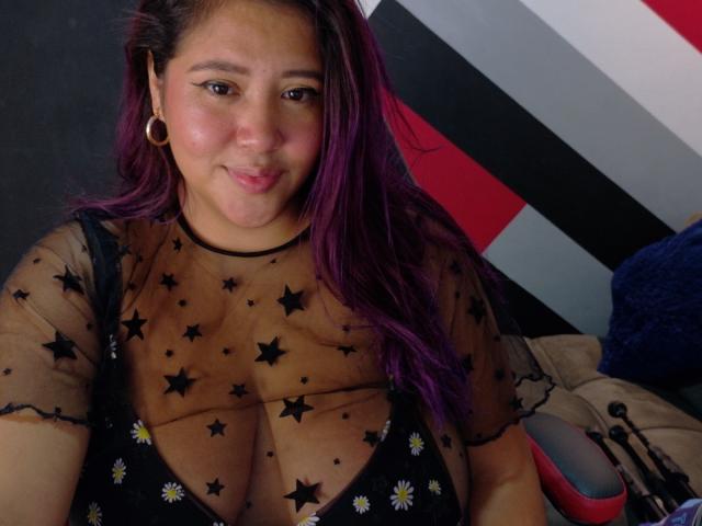 IamAhYours has medium brown skin and purple-dyed hair. She wears a sheer black top with star patterns and small white daisy prints, revealing a deep cleavage. Her black eyeliner is prominent, and she has small gold earrings. She sits on a bed with a gray and red striped wall in the background. The composition is close-up, focusing on her upper body and face. She has a slight, confident smile. The image has a casual, personal feel.