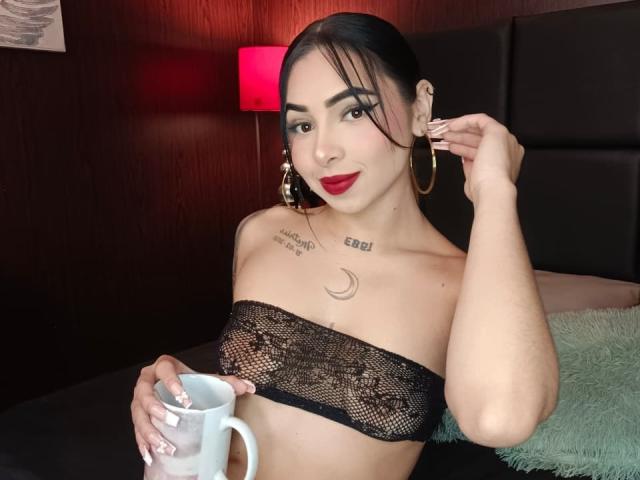 MistressAsshley with dark hair, red lipstick, hoop earrings, black lace tube top, holding a white mug, tattoos on chest and neck, in a dimly lit room.