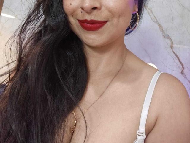 StangelarRivera with long black hair, red lipstick, small mole on her upper lip, white bra straps, gold necklace, and purple earrings, showing her tits and smiling slightly.