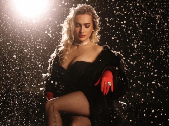 KateOweens with blonde curls, wearing a black dress, red gloves, and fur coat, snowing, showing tits, tattoo on left thigh, glowing light source, focused expression, dark background.